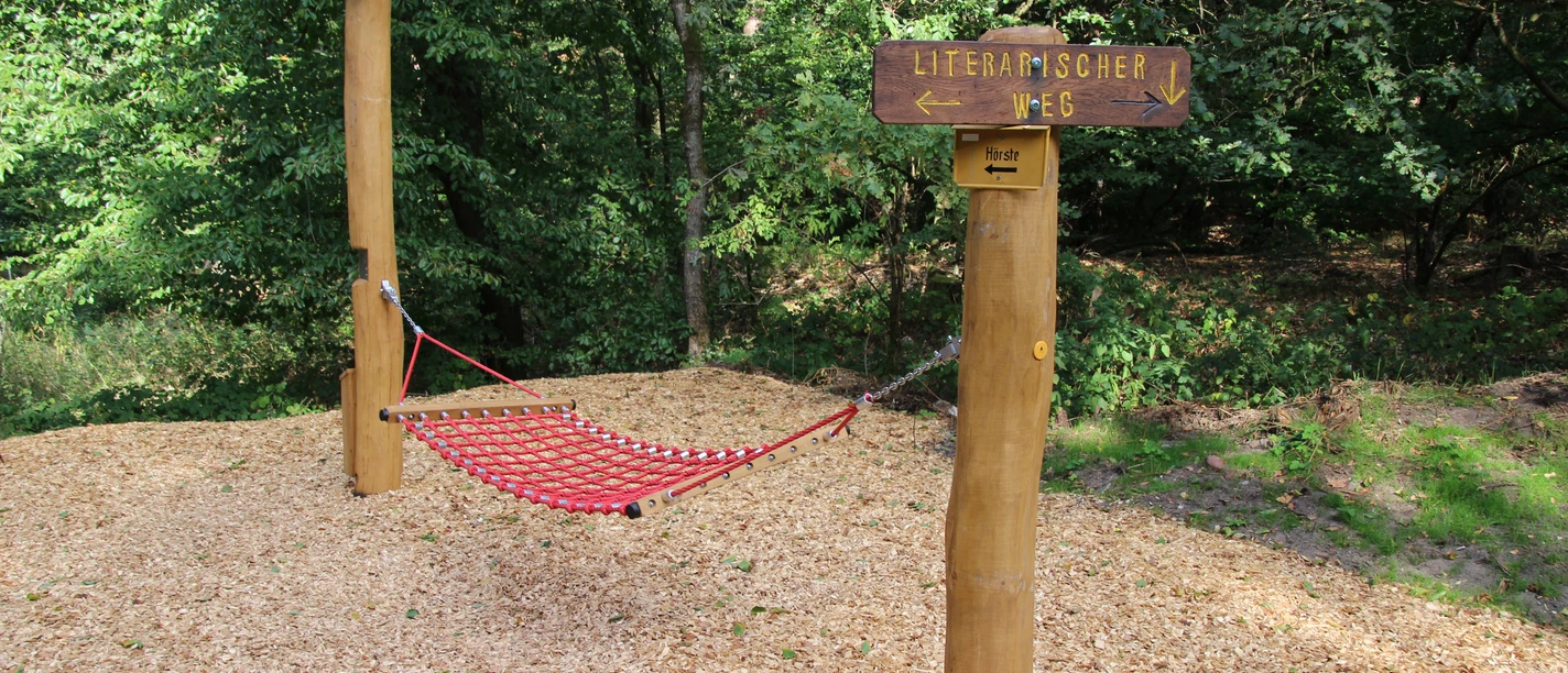 Hängematte Rote Hängematte aus robustem Material im Wald, flankiert von Holzstangen und Wegweiser "Literarischer Weg".
