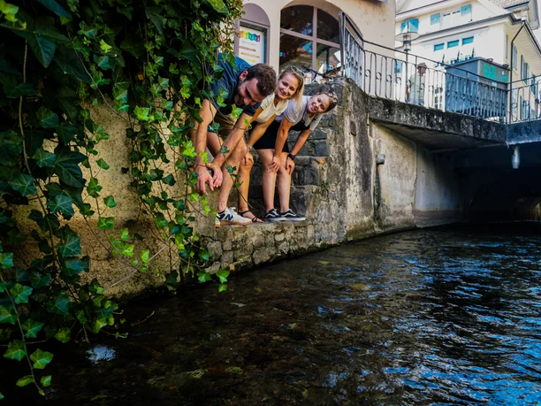 Drei Personen suchen Hinweise an einem Bachufer unter einer Brücke.