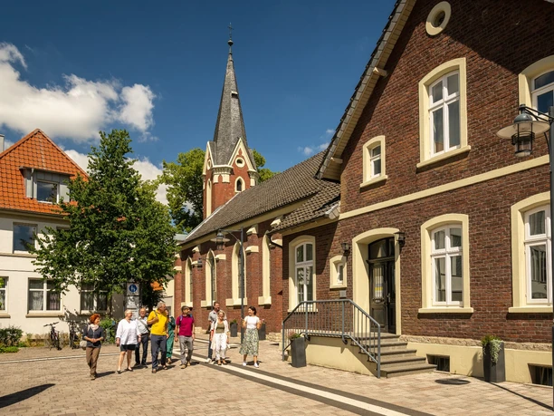 Eine Gruppe von Menschen spaziert über einen sonnigen Platz mit historischen Backsteingebäuden.