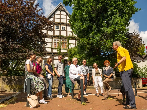 Eine Gruppe von Menschen sitzt und steht vor einem Fachwerkhaus unter blauem Himmel mit Wolken.