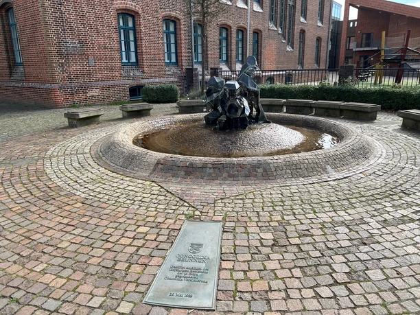 A round stone fountain with a bronze sculpture in the middle of a paved courtyard.