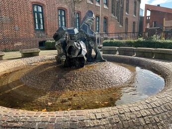 Ein moderner Brunnen mit fließendem Wasser vor einer Backsteinfassade und einigen Sitzgelegenheiten drumherum.
