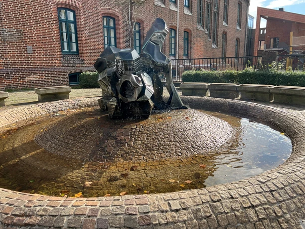 Ein moderner Brunnen mit fließendem Wasser vor einer Backsteinfassade und einigen Sitzgelegenheiten drumherum.