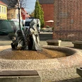 Zwei moderne Zwergenfiguren sprudeln Wasser auf einem gepflasterten zentralen Platz mit Sitzgelegenheiten.