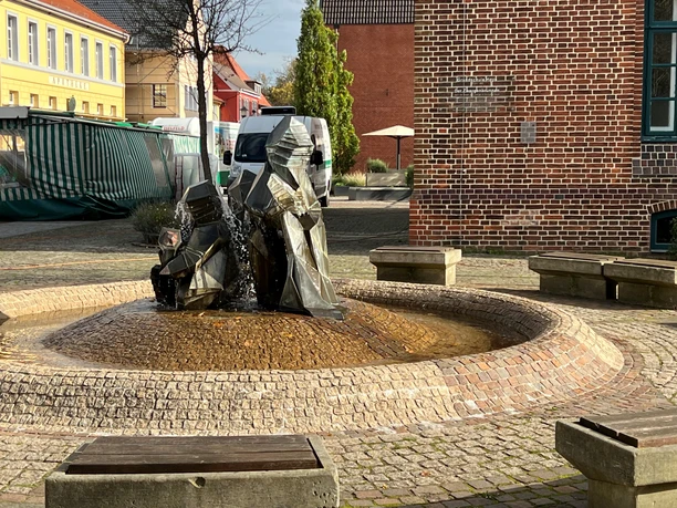 Zwei moderne Zwergenfiguren sprudeln Wasser auf einem gepflasterten zentralen Platz mit Sitzgelegenheiten.