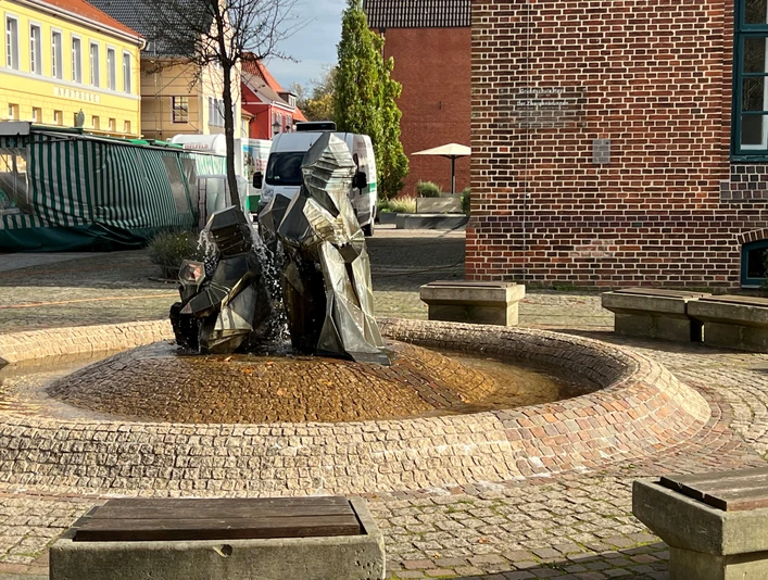 Zwei moderne Zwergenfiguren sprudeln Wasser auf einem gepflasterten zentralen Platz mit Sitzgelegenheiten.