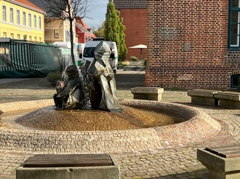 Zwergenbrunnen Zwei moderne Zwergenfiguren sprudeln Wasser auf einem gepflasterten zentralen Platz mit Sitzgelegenheiten.