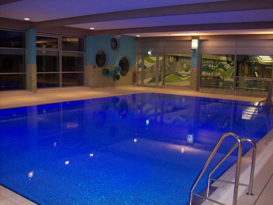 SchwimmWerk Steinhagen Lehrschwimmbecken Hallenbad mit beleuchtetem Innenpool, modernen Glasfronten und türkisfarbener Wandgestaltung.