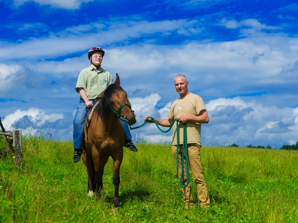 Andershof_Pferd_Wiese Zwei Personen mit einem Pferd auf einer grünen Wiese unter blauem Himmel.