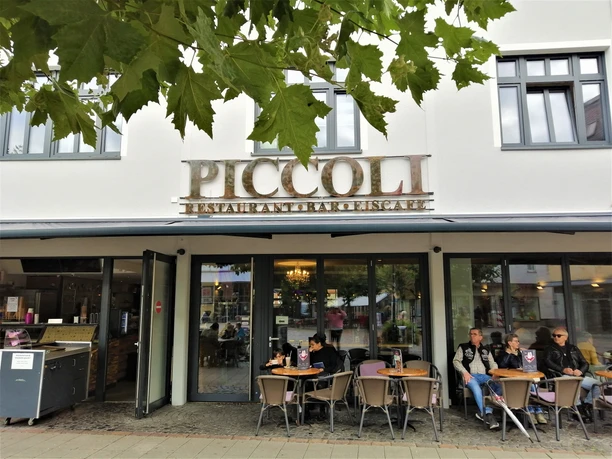 Außenansicht des Restaurants Piccoli mit Gästen, die an Tischen vor großen Fenstern sitzen.