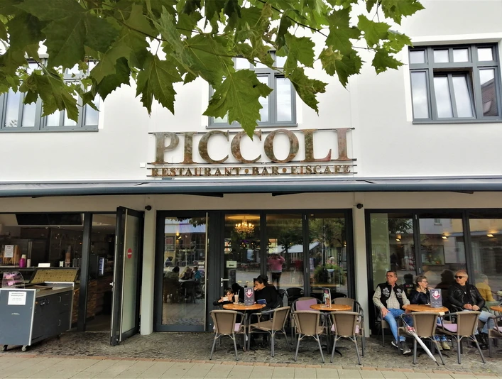 Herford-Piccoli-Aussen-Pro Herford GmbH.jpg Außenansicht des Restaurants Piccoli mit Gästen, die an Tischen vor großen Fenstern sitzen.