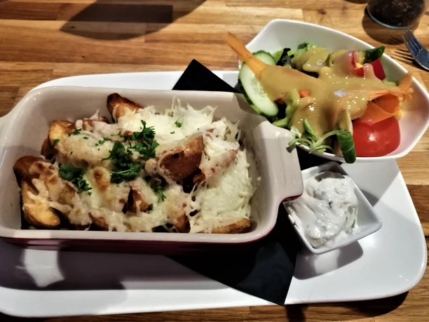 Kartoffelspalten mit geschmolzenem Käse, frische Salatgarnitur und Kräuterdipp auf einem Holztablett.