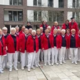 Delme Shanty Singers Große Gesangsgruppe in rot-blauer Kleidung steht lächelnd vor einem modernen Gebäudeensemble.