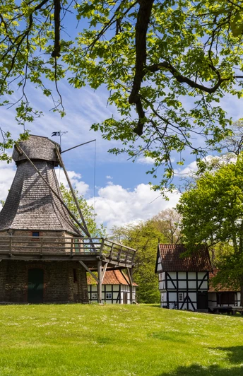 Die historische Mühle Levern mit Schindeldach liegt malerisch in einer grünen Landschaft unter blauem Himmel.