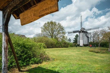 Holz-Bockwindmühle in Landschaft mit grüner Wiese und Leuten, darunter ein Schild mit "Herzlich Willkommen".