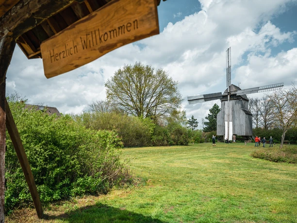 Bockwindmühle Oppenwehe Holz-Bockwindmühle in Landschaft mit grüner Wiese und Leuten, darunter ein Schild mit "Herzlich Willkommen".