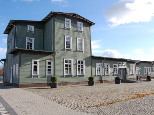 Historisches Bahnhofsgebäude aus Holz mit grüner Fassade und weißen Fenstern im Sonnenschein.