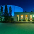 Ein beleuchtetes Gebäude bei Nacht mit blauer Kuppel im Hintergrund und Eingangsbereich im Vordergrund.