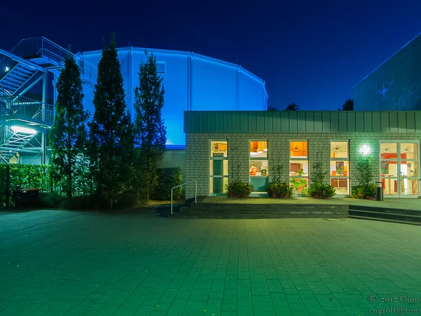 Ein beleuchtetes Gebäude bei Nacht mit blauer Kuppel im Hintergrund und Eingangsbereich im Vordergrund.