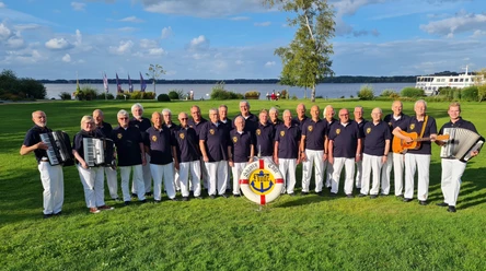 Shanty-Chor Hude Eine Gruppe männlicher Sänger des Shanty-Chors Hude steht in einer Reihe im Kurpark mit einem Akkordeonspieler links und einem Gitarristen rechts. Im Hintergrund ist das Zwischenahner Meer und ein blauer Himmel zu sehen.