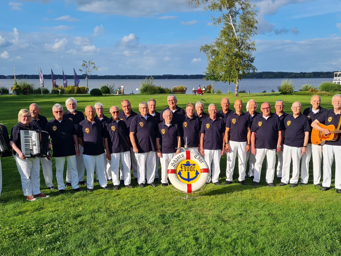 Shanty-Chor Hude Eine Gruppe männlicher Sänger des Shanty-Chors Hude steht in einer Reihe im Kurpark mit einem Akkordeonspieler links und einem Gitarristen rechts. Im Hintergrund ist das Zwischenahner Meer und ein blauer Himmel zu sehen.