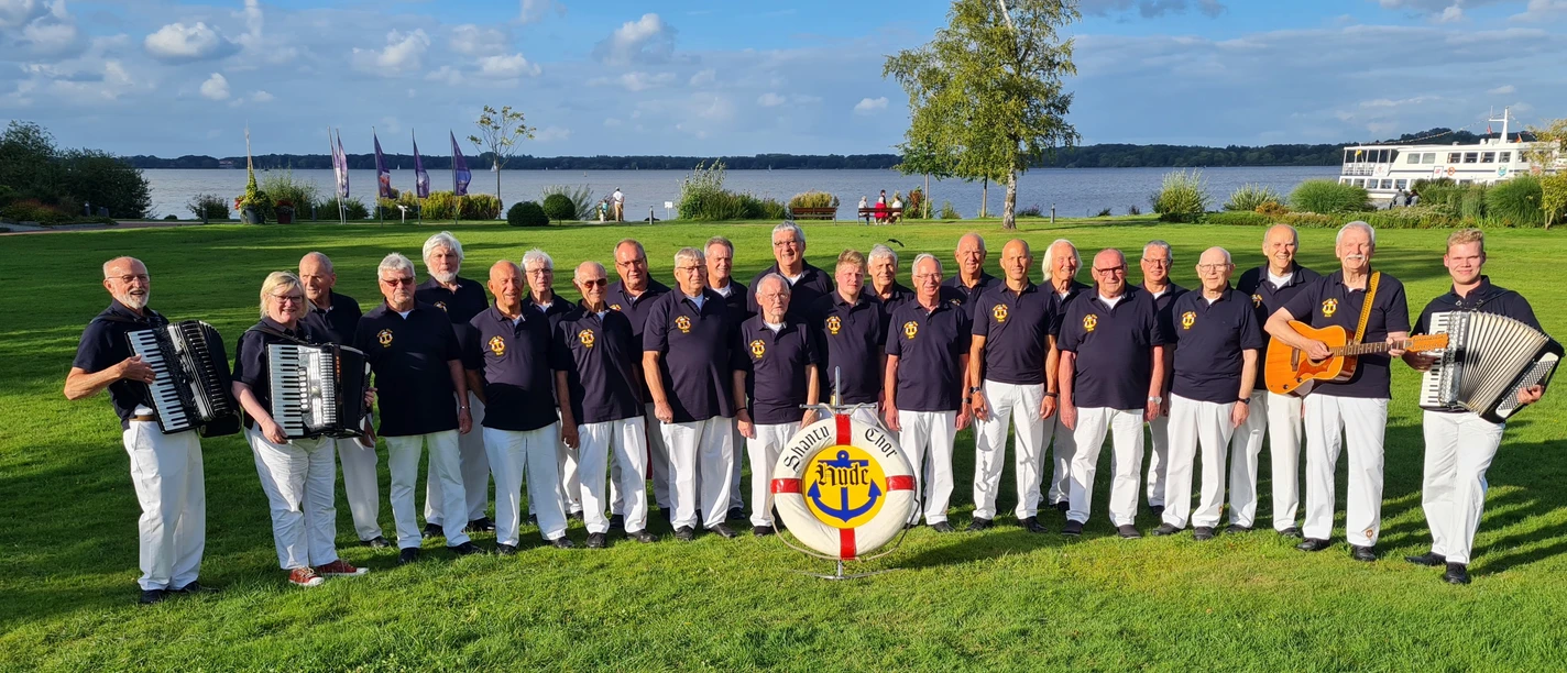 Shanty-Chor Hude Eine Gruppe männlicher Sänger des Shanty-Chors Hude steht in einer Reihe im Kurpark mit einem Akkordeonspieler links und einem Gitarristen rechts. Im Hintergrund ist das Zwischenahner Meer und ein blauer Himmel zu sehen.