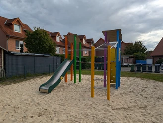 Ein bunter Spielplatz mit Klettergerüst und Rutsche, umgeben von Sand und Wohnhäusern im Hintergrund.