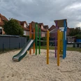 Ein bunter Spielplatz mit Klettergerüst und Rutsche, umgeben von Sand und Wohnhäusern im Hintergrund.
