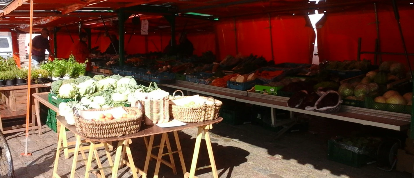 Stand auf dem Aper Wochenmarkt Ein Marktstand mit frischen Gemüsekisten unter orangefarbenen Planen auf einem gepflasterten Platz.