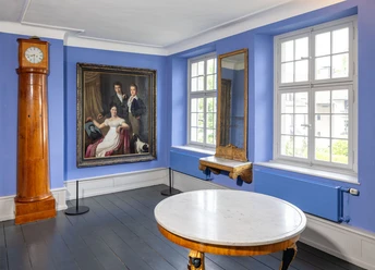 Die Brügelmanns Cromford Ein lichtdurchfluteter Raum mit blauen Wänden, Holzuhr, Gemälde, Spiegel und einem runden Tisch.