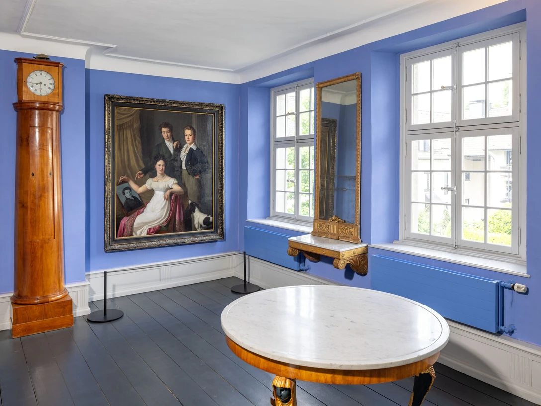 Die Brügelmanns Cromford Ein lichtdurchfluteter Raum mit blauen Wänden, Holzuhr, Gemälde, Spiegel und einem runden Tisch.