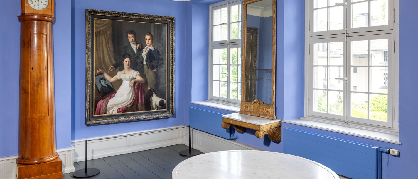 Die Brügelmanns Cromford Ein lichtdurchfluteter Raum mit blauen Wänden, Holzuhr, Gemälde, Spiegel und einem runden Tisch.