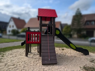 Holzspielplatz mit Rutsche, Kletterturm und Sandboden in einer Wohngegend mit Häusern im Hintergrund.