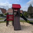 Holzspielplatz mit Rutsche, Kletterturm und Sandboden in einer Wohngegend mit Häusern im Hintergrund.