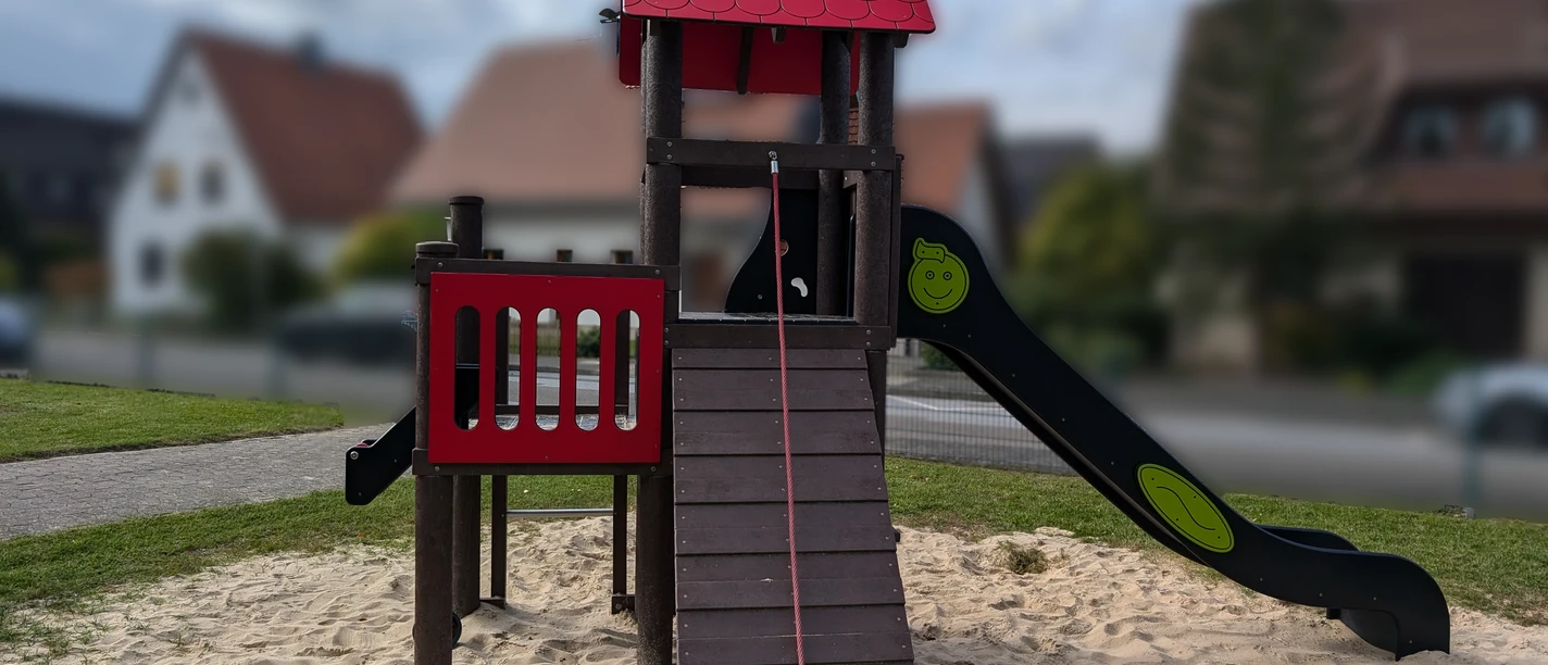 Holzspielplatz mit Rutsche, Kletterturm und Sandboden in einer Wohngegend mit Häusern im Hintergrund.