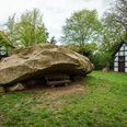 Großer Findling in Wiese bei Fachwerkhäusern, gerahmt von Bäumen, parkähnliche Umgebung.