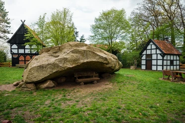 Großer Stein Tonnenheide Großer Findling in Wiese bei Fachwerkhäusern, gerahmt von Bäumen, parkähnliche Umgebung.