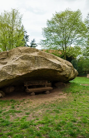 Großer Stein Tonnenheide Großer Findling in Wiese bei Fachwerkhäusern, gerahmt von Bäumen, parkähnliche Umgebung.