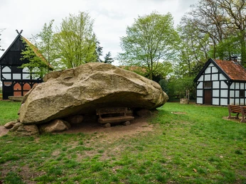 Großer Stein Tonnenheide Großer Findling in Wiese bei Fachwerkhäusern, gerahmt von Bäumen, parkähnliche Umgebung.