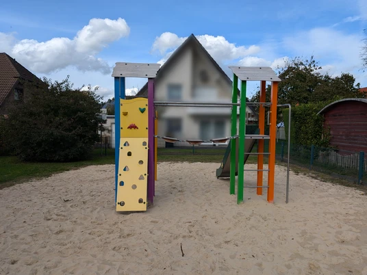 Spielplatz mit Kletterturm, Rutsche und Kletterwand auf Sandboden, umgeben von Büschen und Häusern.