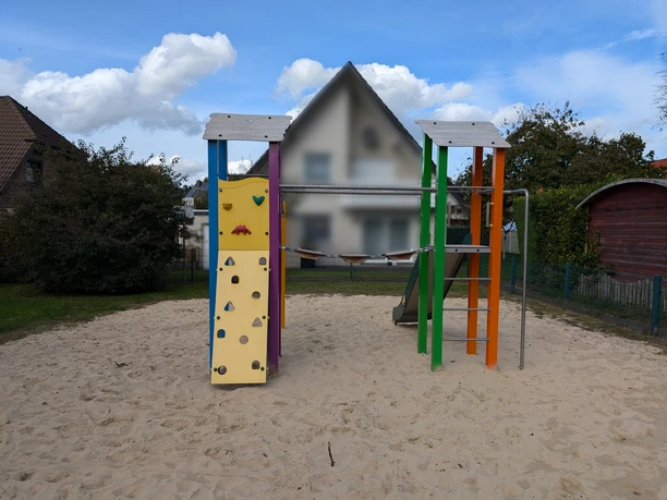 Verl-SpielplatzAnnaburgerstraße-Teutoburger-Wald-StadtVerl Spielplatz mit Kletterturm, Rutsche und Kletterwand auf Sandboden, umgeben von Büschen und Häusern.