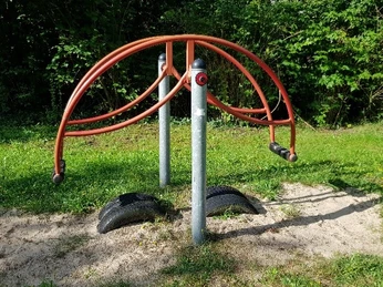 Ein moderner Spielplatz mit Metallgerüst zum Klettern steht auf sandigem Boden, umgeben von Gras.