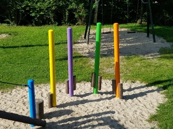 Bunte Balancierstangen in einem Sandbereich, umgeben von grüner Wiese und Bäumen, mit Schaukeln im Hintergrund.