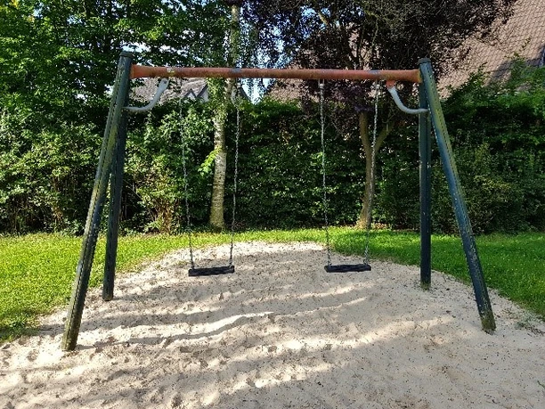 Zwei Schaukeln auf grünem Spielplatz, umgeben von Bäumen und Sandboden bei sonnigem Wetter.