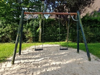 2025_Wilhelm-Busch-Straße_Doppelschaukel Zwei Schaukeln auf grünem Spielplatz, umgeben von Bäumen und Sandboden bei sonnigem Wetter.