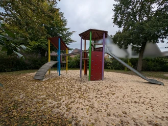 Ein moderner Spielplatz mit Klettergerüst, Rutsche und Wipptieren, umgeben von herbstlichem Laub.