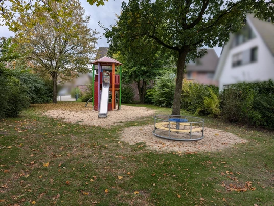 Ein Spielplatz mit Kletterturm, Rutsche und Karussell, umgeben von Bäumen und Häusern im Hintergrund.
