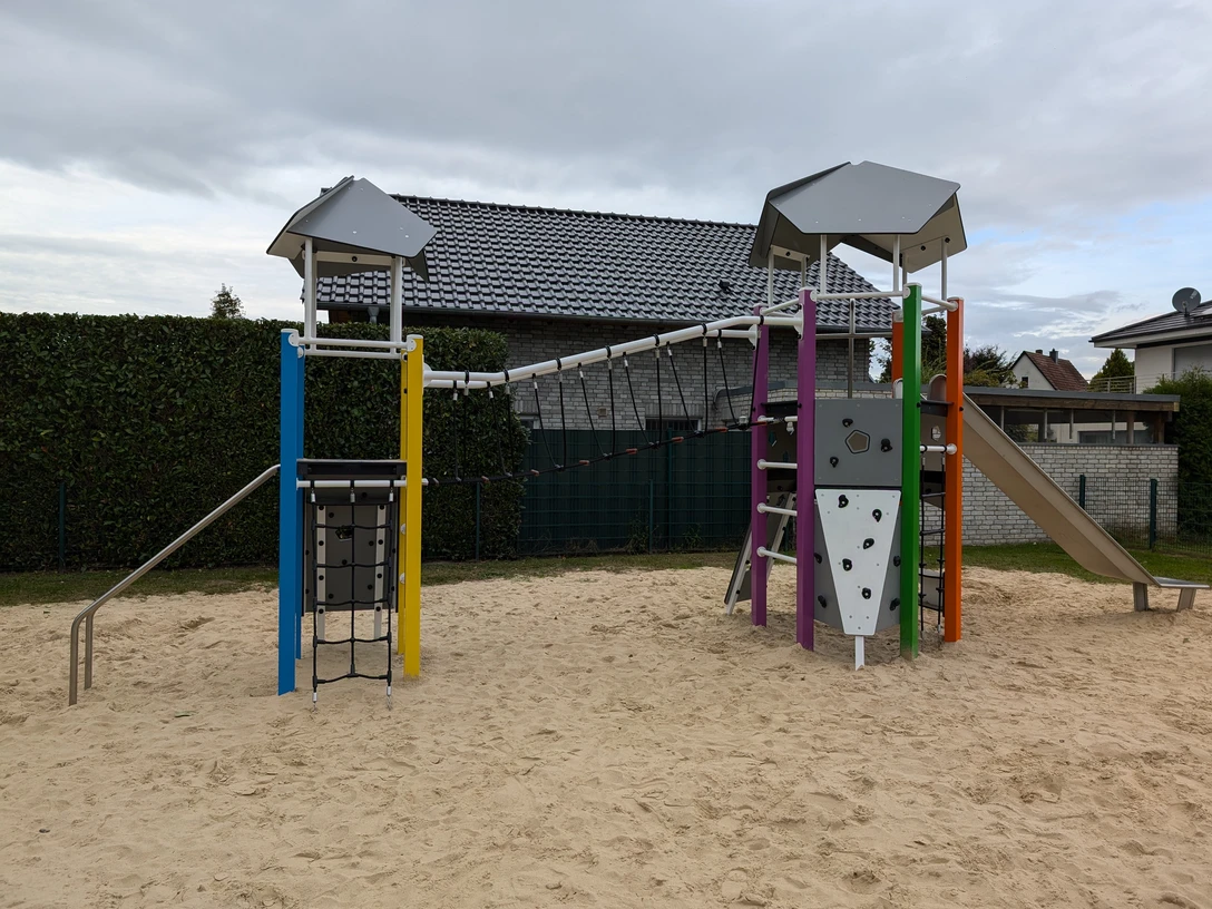 Spielplatz Varusweg Moderner Spielplatz mit Klettergerüst, Rutsche und Sandfläche, umgeben von Häusern und Hecken.