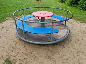 Karussell Rundes Karussell auf einem Spielplatz mit blauen Sitzen und einem roten Tisch in sandiger Umgebung.