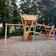 Kombispielgerät Ein moderner Spielplatz aus Holz, umgeben von Bäumen, bietet Kletter- und Balanciermöglichkeiten.